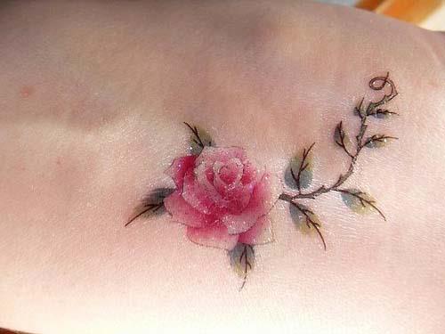 An artistic tattoo of a pink rose flower. The stem of the rose has thorns, and the leaves have prickly tips