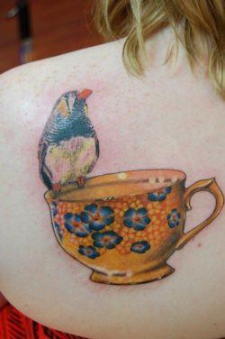 A tattoo design of a yellow tea cup with blue flowers and a little bird decorates this girls shoulder