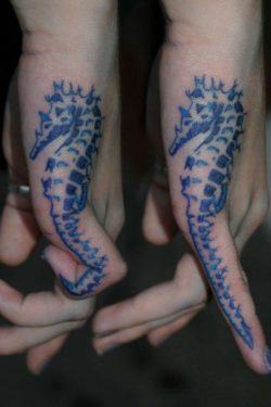 A blue ink tattoo of a seahorse on the side of the hand. When the tattoo owner curls her finger, the tail of the seahorse curls too