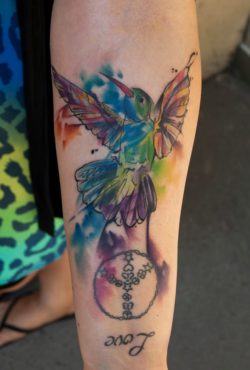 Versatile tattoo artist Csiga creates a watercolor tattoo of a hummingbird with a peace sign and the word Love