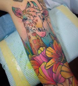 A baby giraffe peeks out over a bed of lily flowers in this colorful watercolor tattoo by Katie Shocrylas