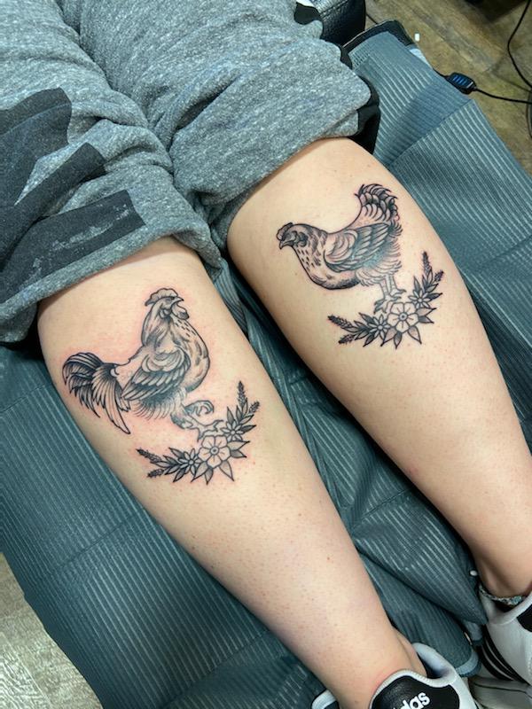 This timeless illustration style tattoo of a rooster and his hen is by tattoo artist Donna Phillips. The illustrative style is of the type that might have been used in picture books from the early twentieth century, and reflects the traditional nature of the relationship between a rooster and his hen. A comparison is drawn between a rooster and his hen and the traditional relationship between a husband and his wife. The artist has cleverly placed the tattoos on her client's calves, giving each bird their own, matching canvas.